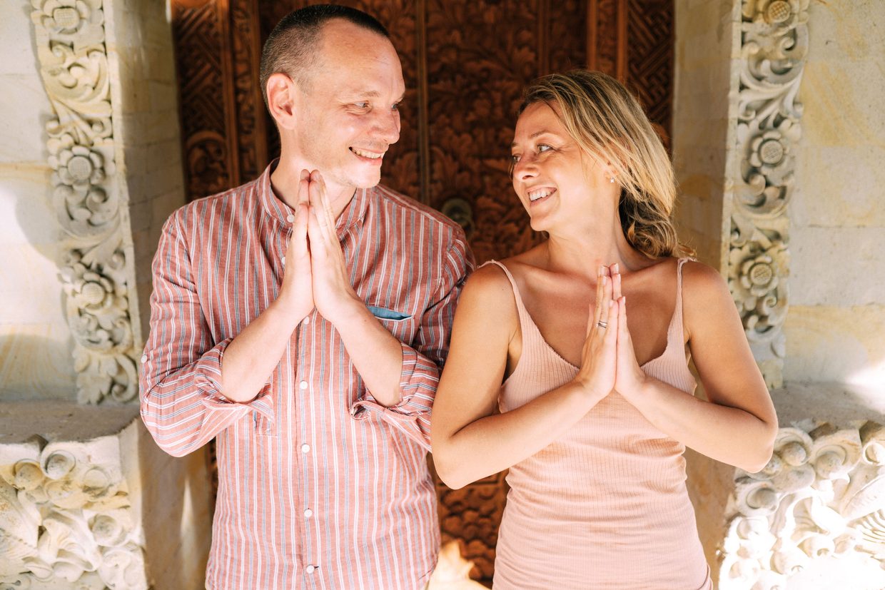 Two people press their palms together in a wai greeting before a carved doorway
