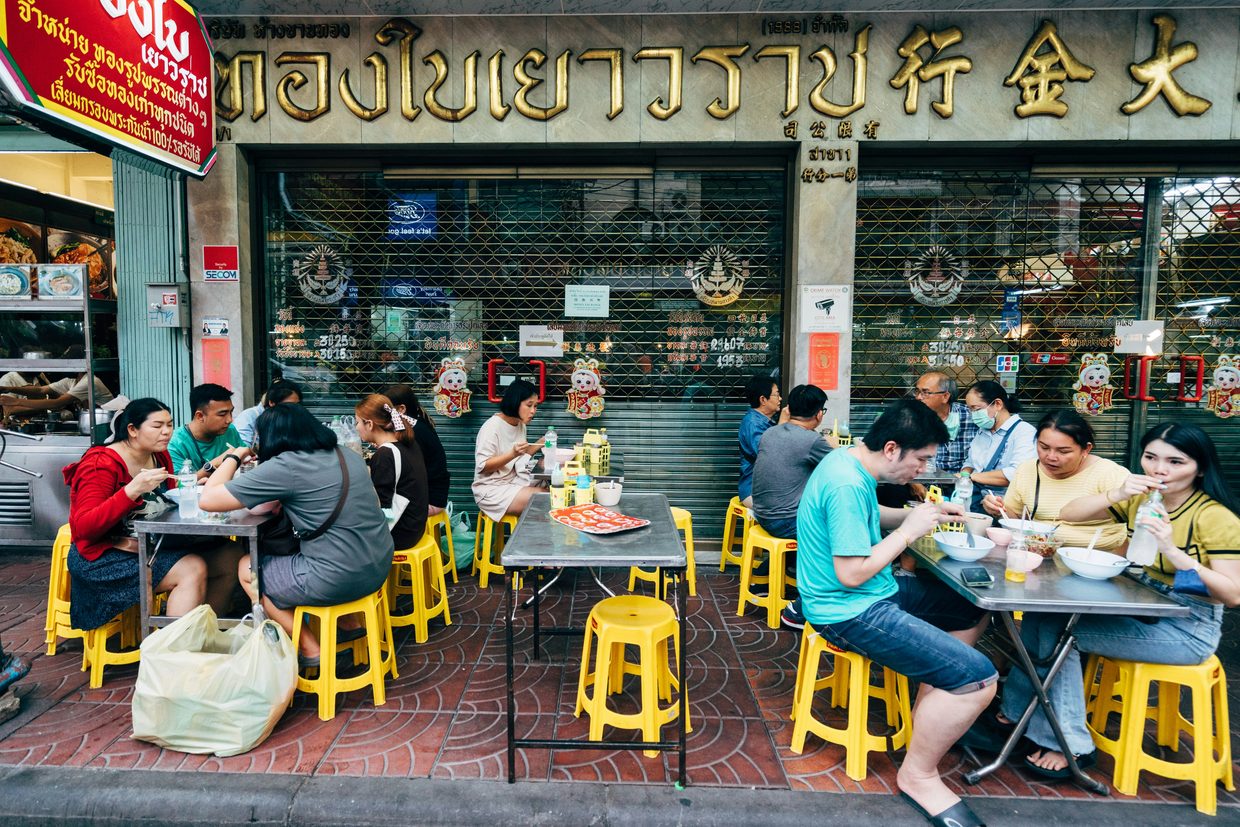 Diners on yellow stools eating noodles at a street stall in Bangkok's Chinatown
