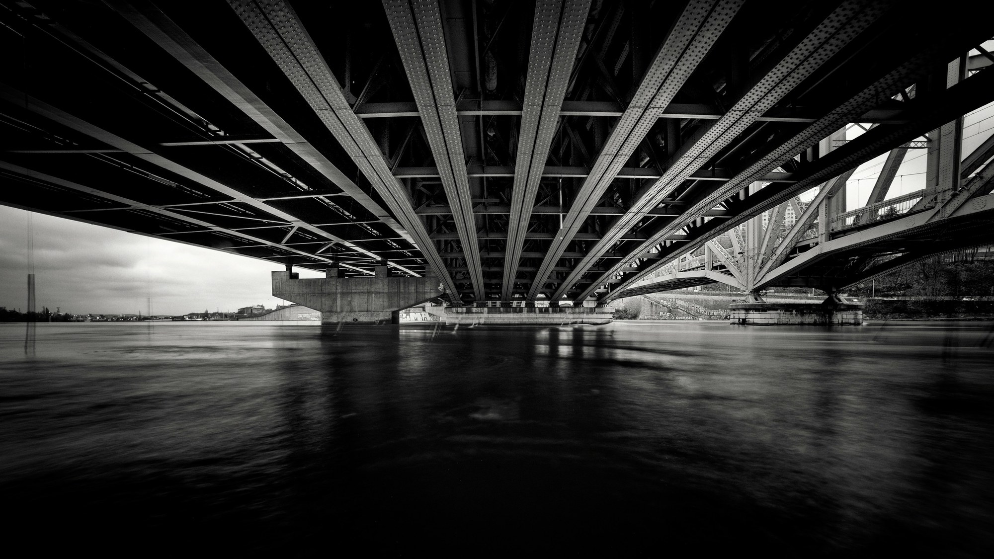 Water under the bridge &mdash; black-and-white view from beneath a steel bridge, its girders converging over dark, moving water